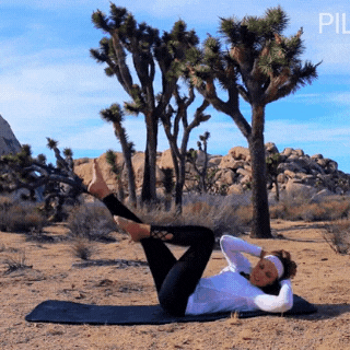 Lisa performs a Pilates exercise on sandy terrain with a desert landscape in the background.
