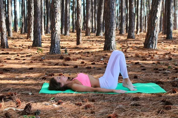 Lisa from Pilates by Lisa is demonstrating a Pilates exercise outdoors on a yoga mat in a woodland setting.